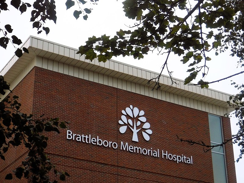 The exterior of Brattleboro Memorial Hospital, featuring its name and logo on a brick building, with tree branches partially framing the view.