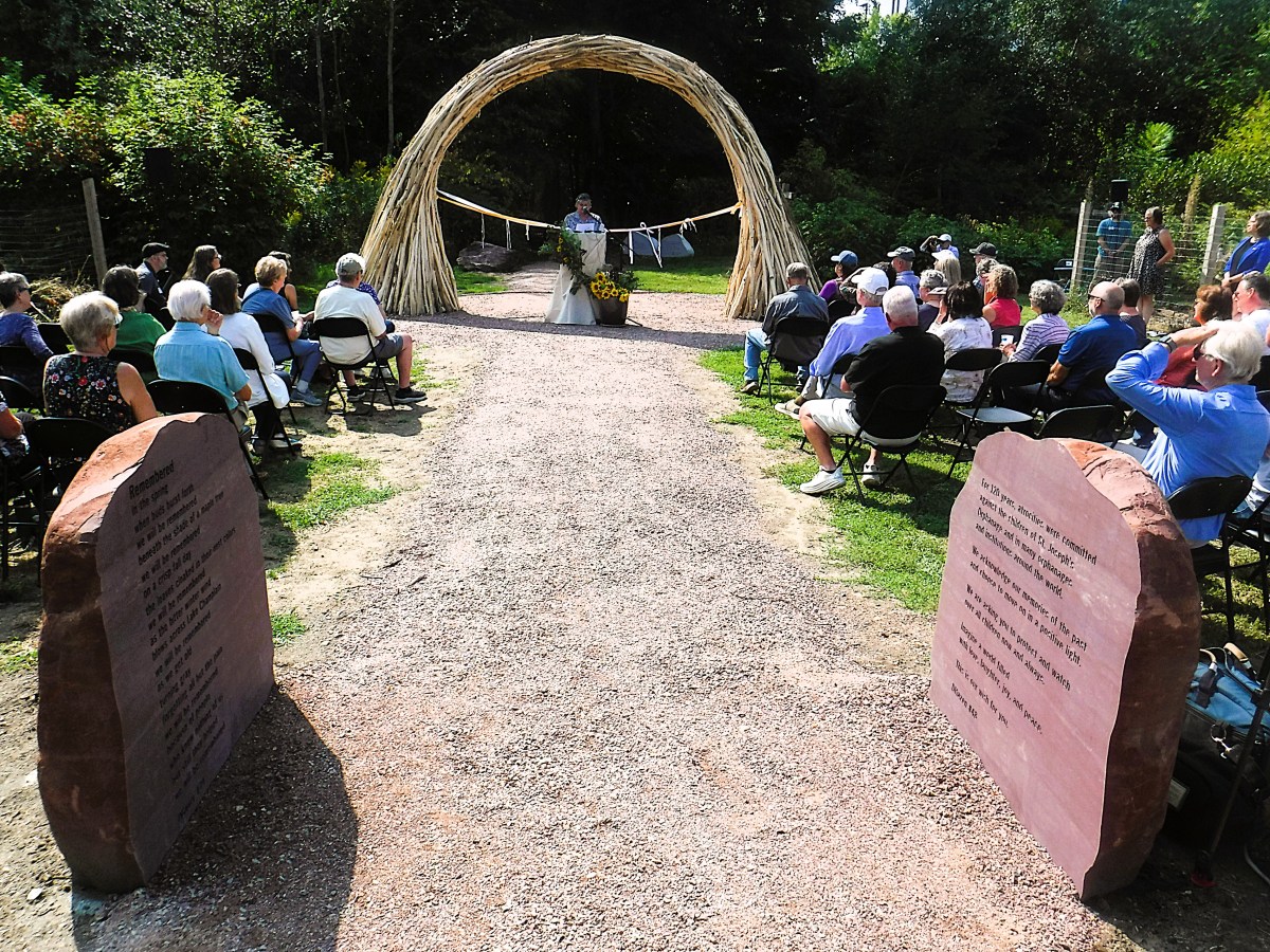 A group of people sit facing a speaker at an outdoor event, gathered under a woven archway with two large engraved stones on either side of the pathway.