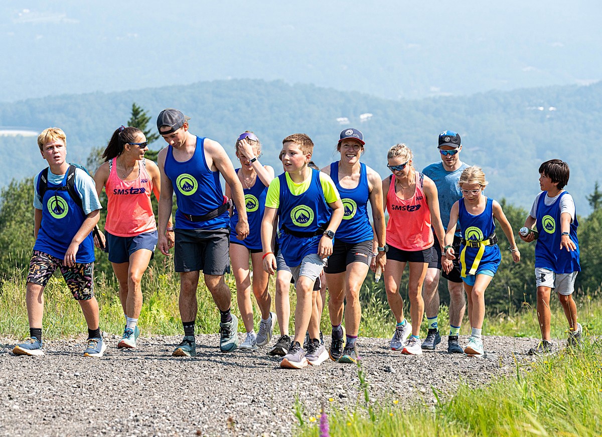 A group of people in athletic clothing hike together up a gravel path, with grassy hills and trees visible in the background.