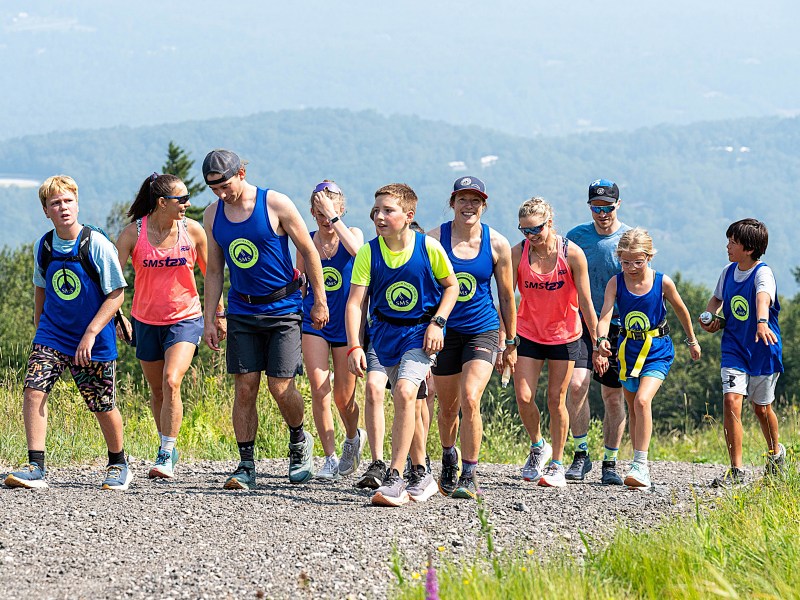 A group of people in athletic clothing hike together up a gravel path, with grassy hills and trees visible in the background.