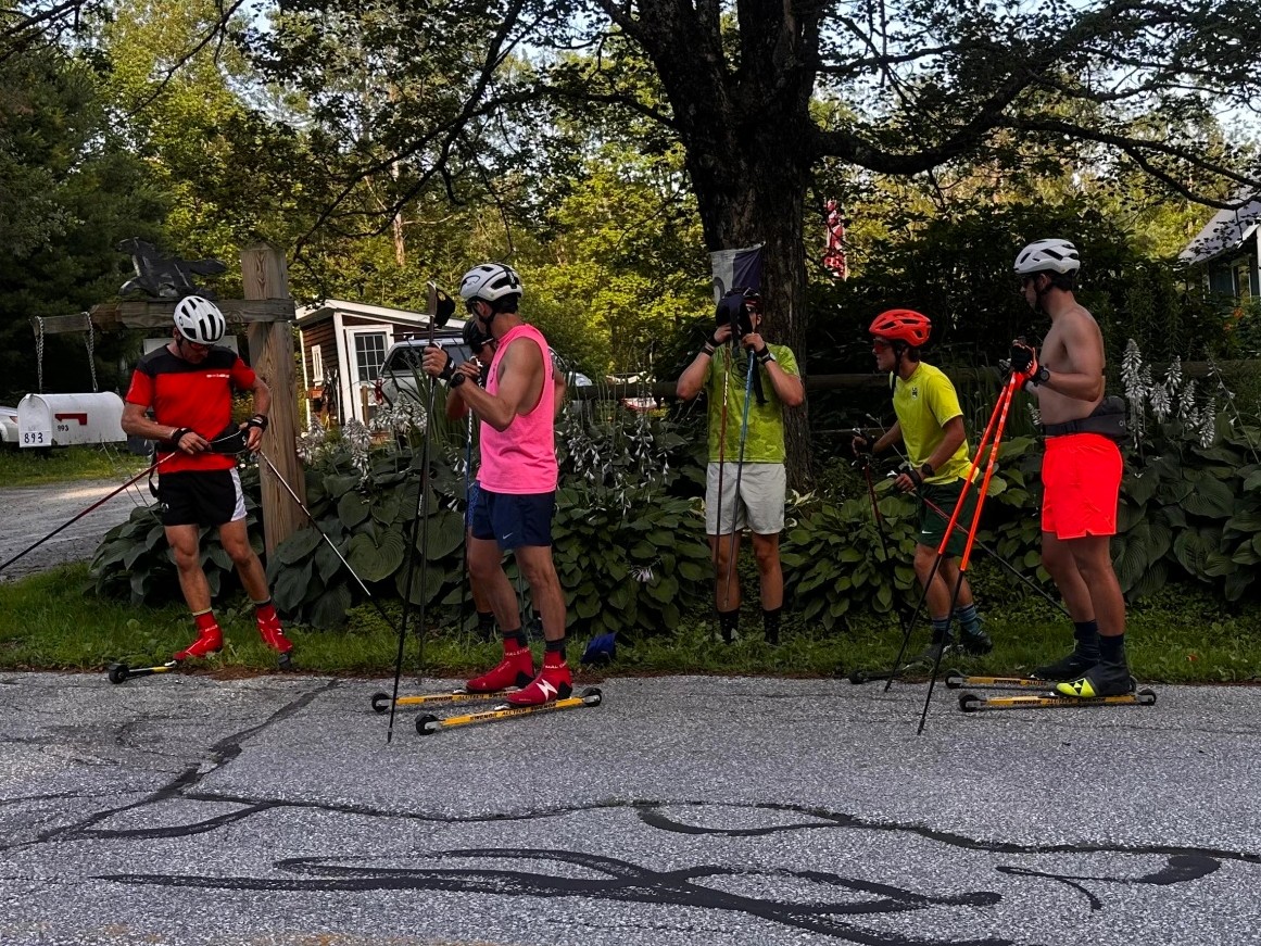 Five people wearing helmets and brightly colored athletic clothing stand on roller skis with poles on a residential street, preparing or taking a break near greenery and mailboxes.