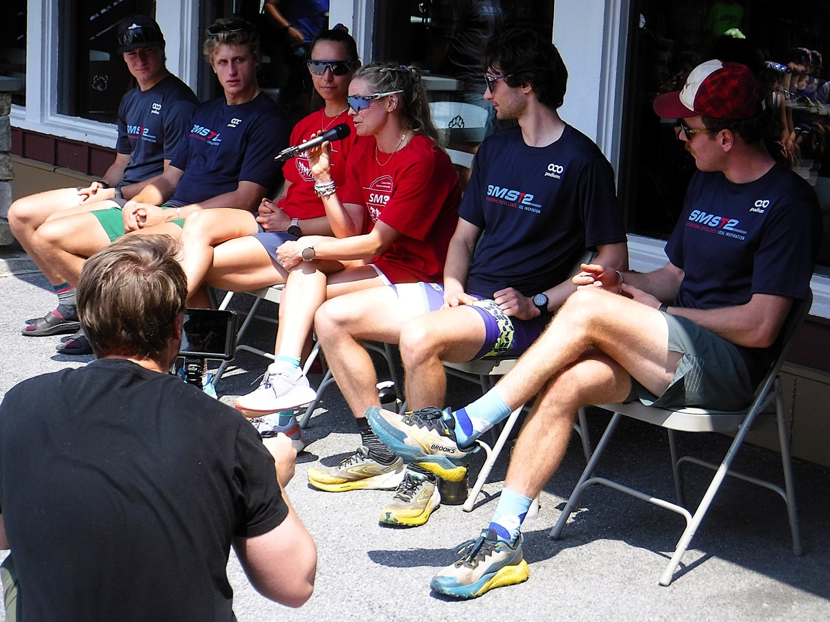 A group of six people sit in chairs outside, with one person speaking into a microphone. Another person kneels in front, filming or taking a photo.