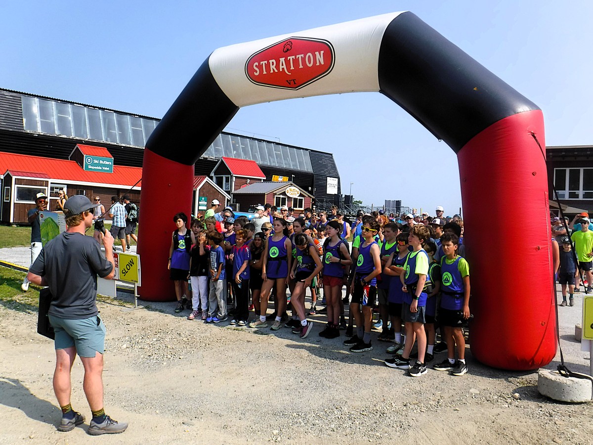 A group of children and teens stand at the starting line under an inflatable arch labeled "Stratton," while a person in shorts and a hat speaks to them.