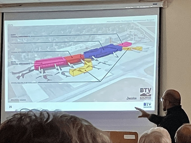 A presenter points to a projected diagram showing a color-coded expansion plan for an airport terminal with labeled sections.