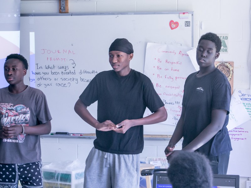 Three teenage boys stand in front of a classroom whiteboard with handwritten notes and posters about respect and race visible behind them.