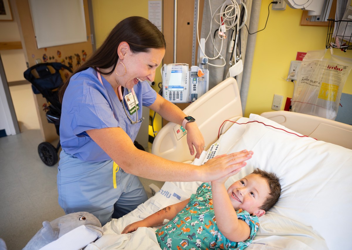 A nurse in scrubs gives a high-five to a smiling child lying in a hospital bed, with medical equipment visible in the background.