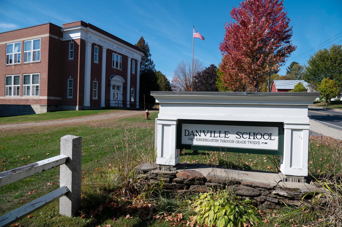 A brick school building with white columns and an American flag stands behind a sign reading "Danville School Pre-Kindergarten through Grade Twelve" on a sunny day.