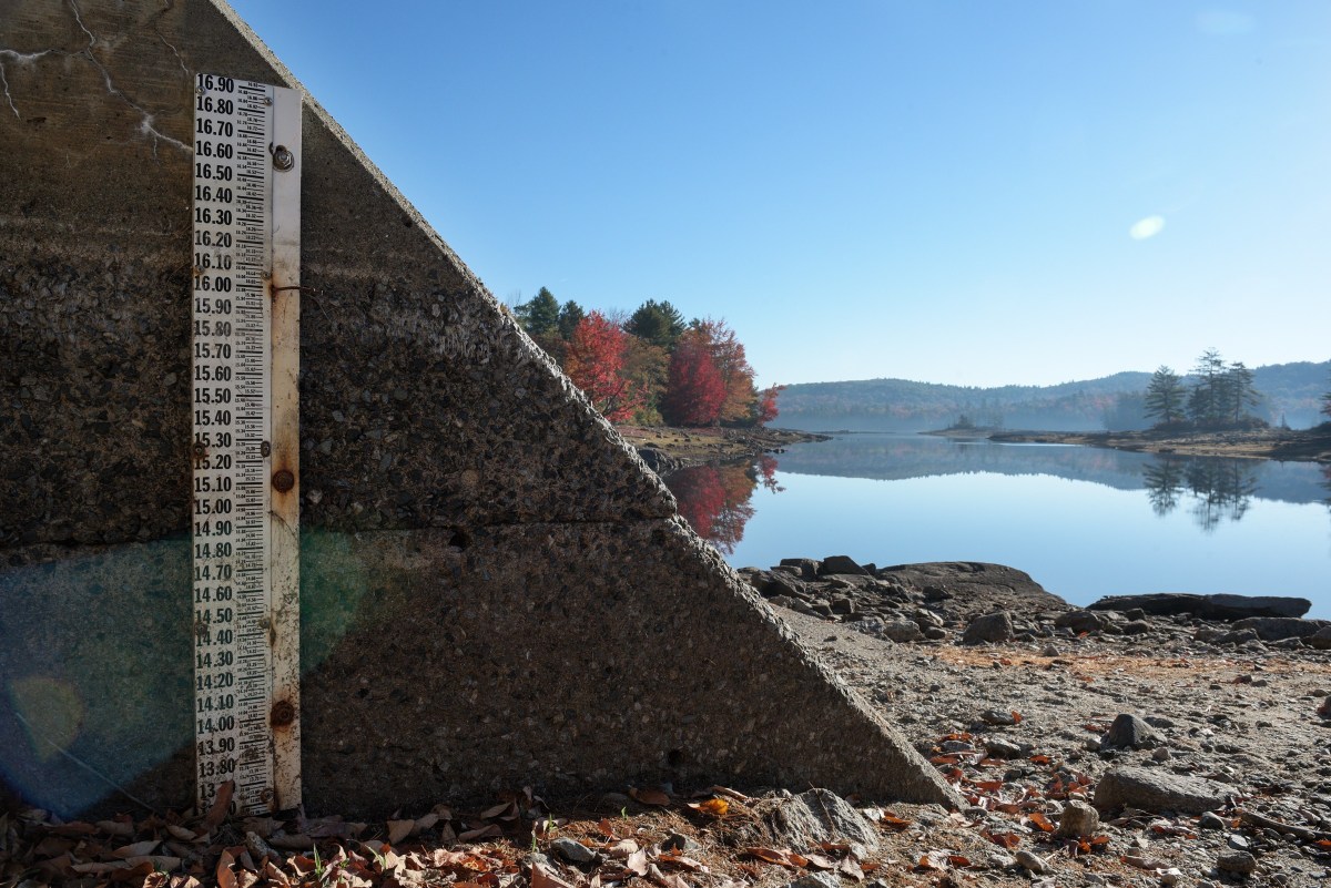 A concrete water level gauge marked with measurements stands beside a calm lake with autumn trees in the distance under a clear blue sky.