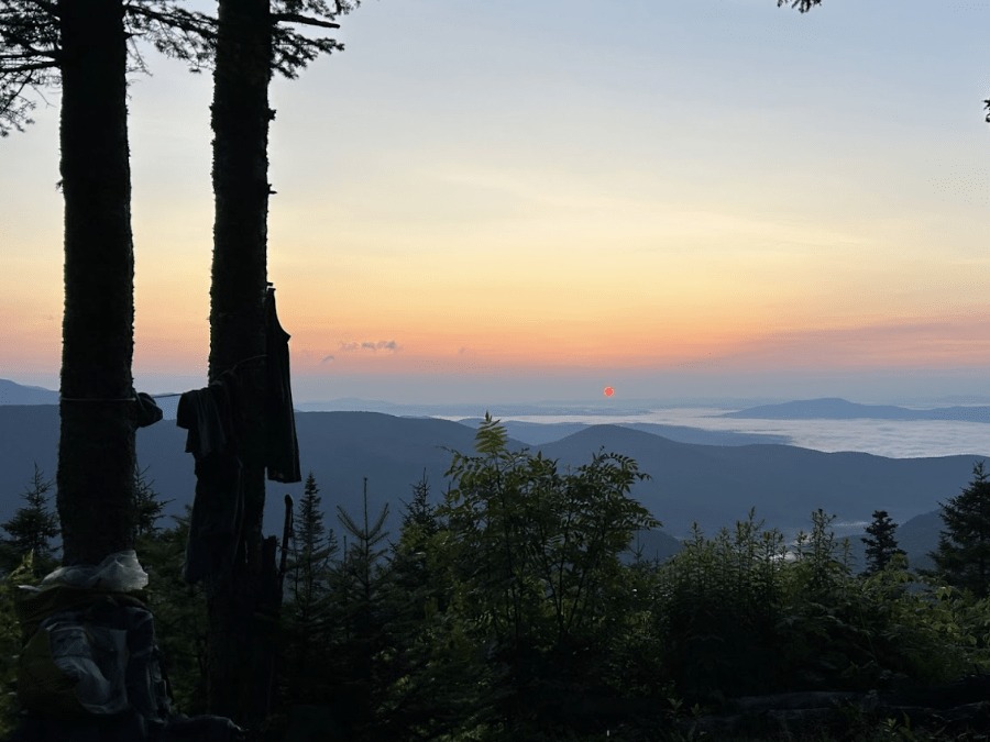 A sunrise view over distant mountains and a valley filled with fog, framed by trees in the foreground with hiking gear hanging on a branch.