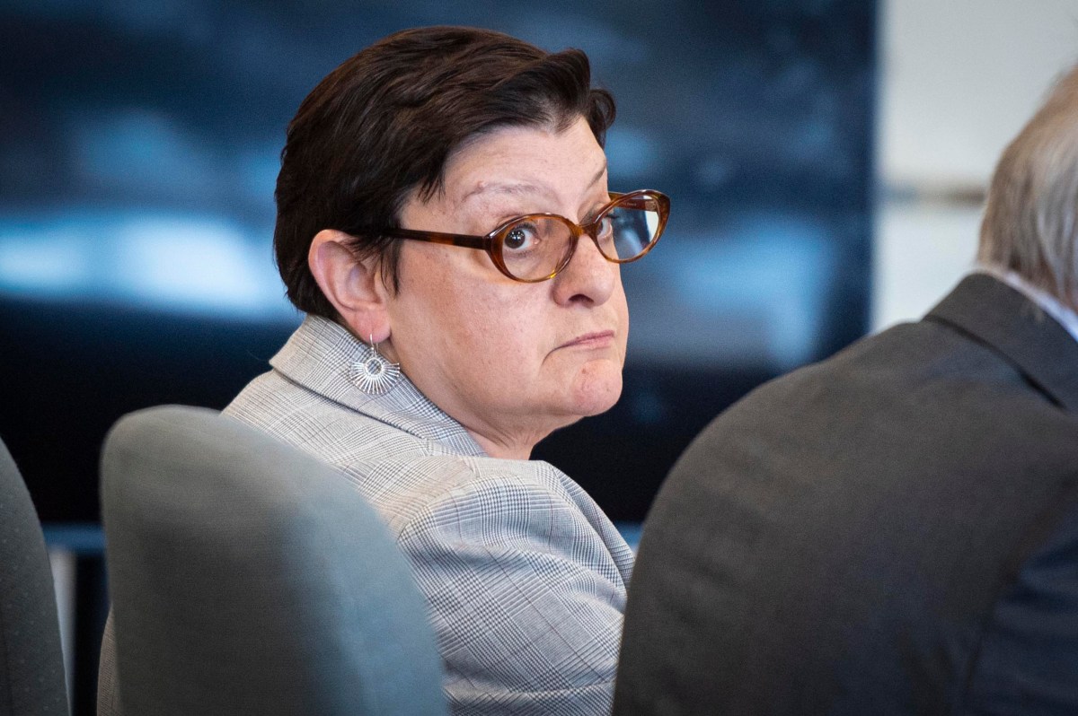 A woman with short dark hair and glasses wearing a gray blazer is seated indoors, looking over her shoulder toward the camera.