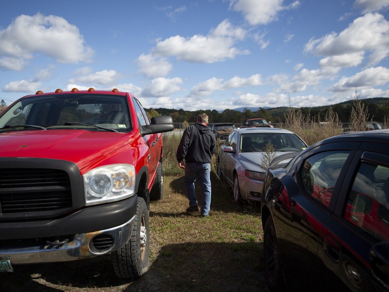 A person walks between parked vehicles, including a red pickup truck, in a grassy outdoor area under a partly cloudy sky.