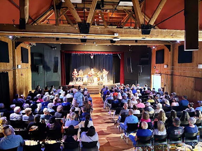 A band performs on stage in front of an audience seated in rows inside a wooden, high-ceilinged venue.