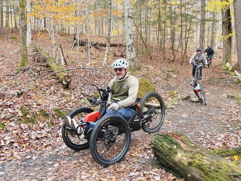 A person riding an off-road adaptive handcycle on a forest trail, followed by two cyclists on standard mountain bikes amid autumn foliage.