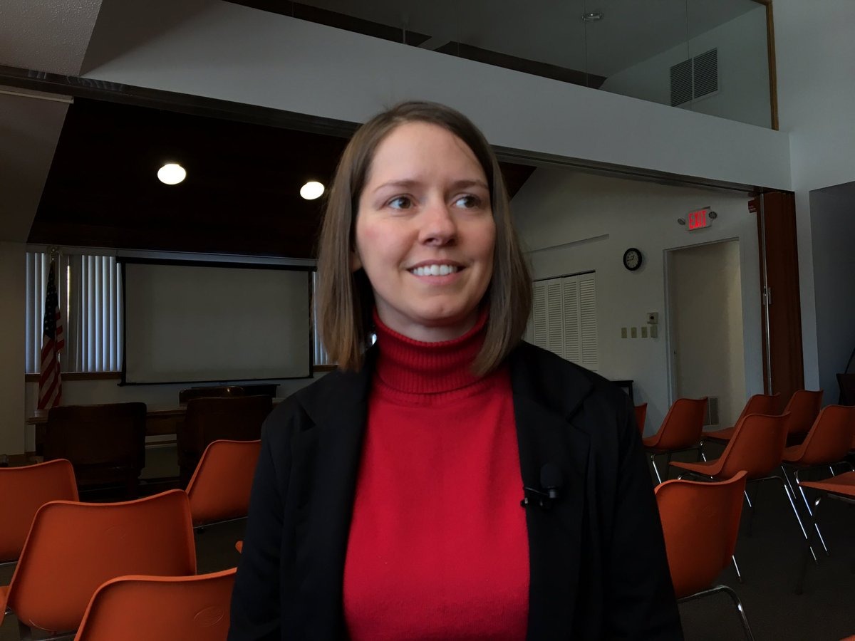 A woman in a red turtleneck and black jacket stands in a room with orange chairs and large windows.