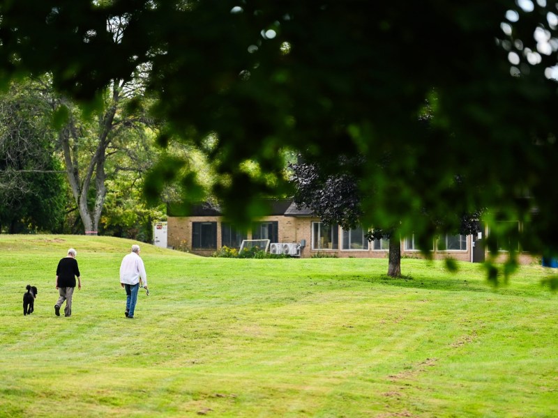 Two people and a black dog walk across a grassy lawn toward a house, with trees and greenery in the background.