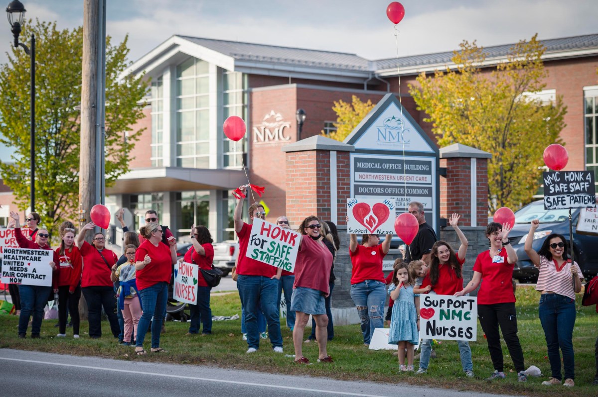 A group of people wearing red hold signs and balloons outside a medical office building, advocating for nurse staffing and support.