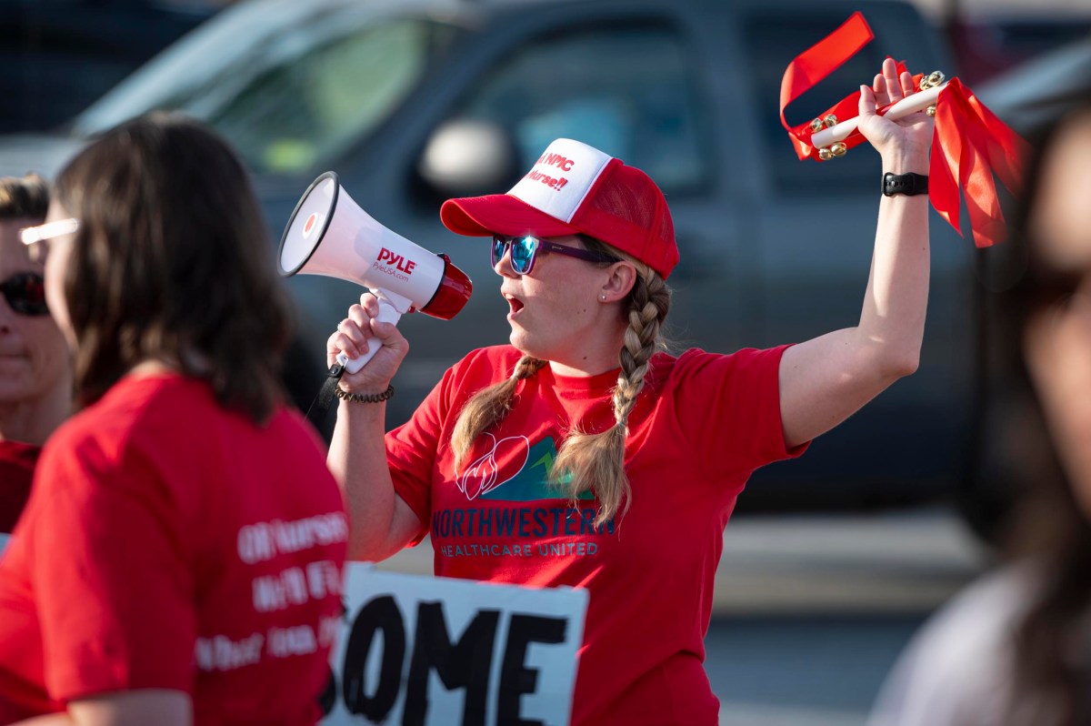 A woman in a red shirt and cap uses a megaphone and holds red ribbons while participating in an outdoor event with other people.