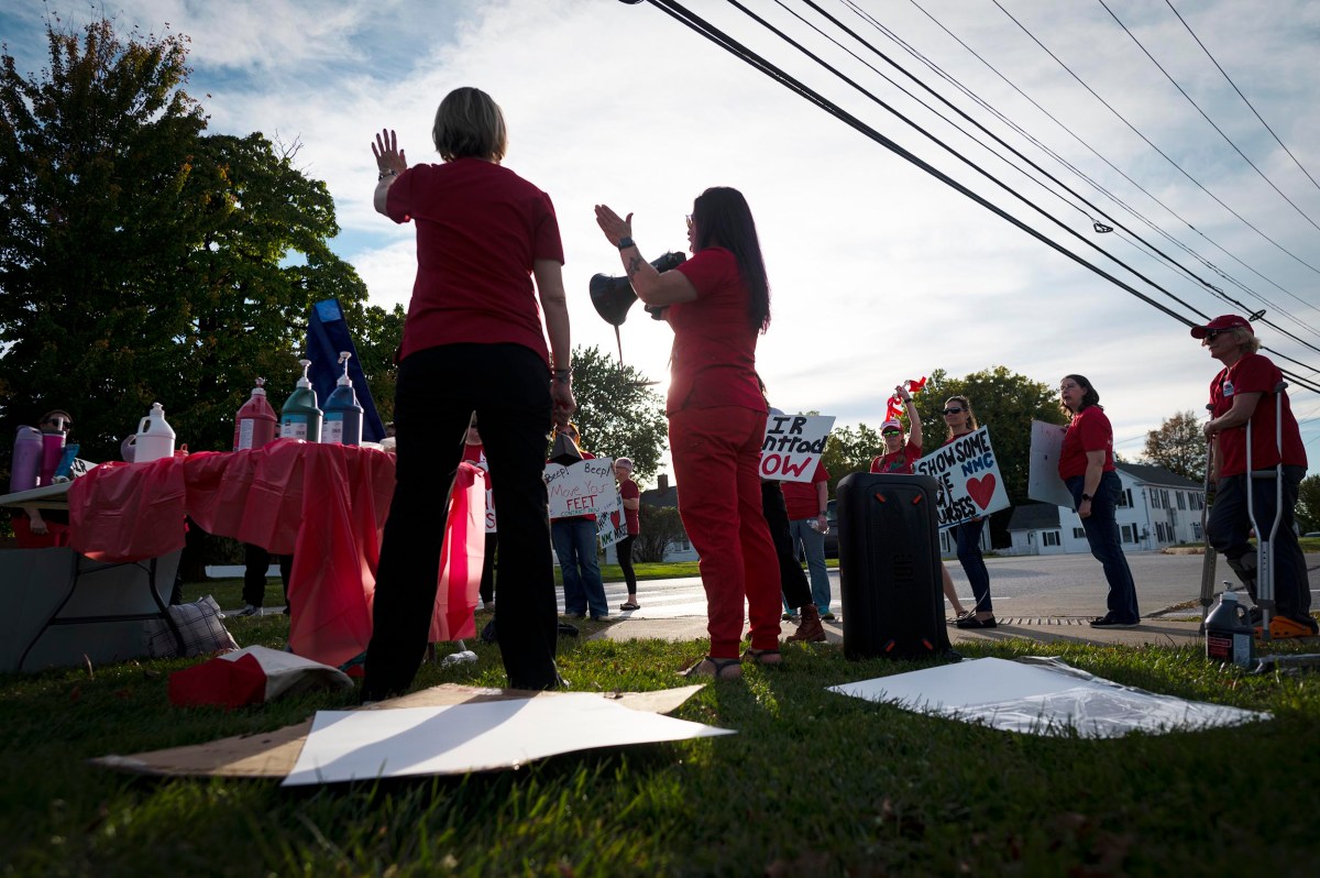 A group of people wearing red clothing hold signs and gather on a sidewalk near a table with supplies, participating in a protest or demonstration on a sunny day.