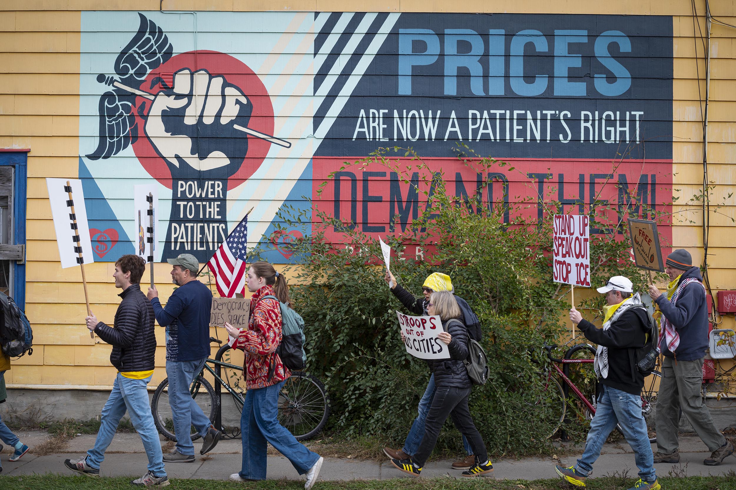 A group of people holding protest signs walks past a mural that reads, “PRICES ARE NOW A PATIENT’S RIGHT. POWER TO THE PATIENTS. DEMAND THEM!”.