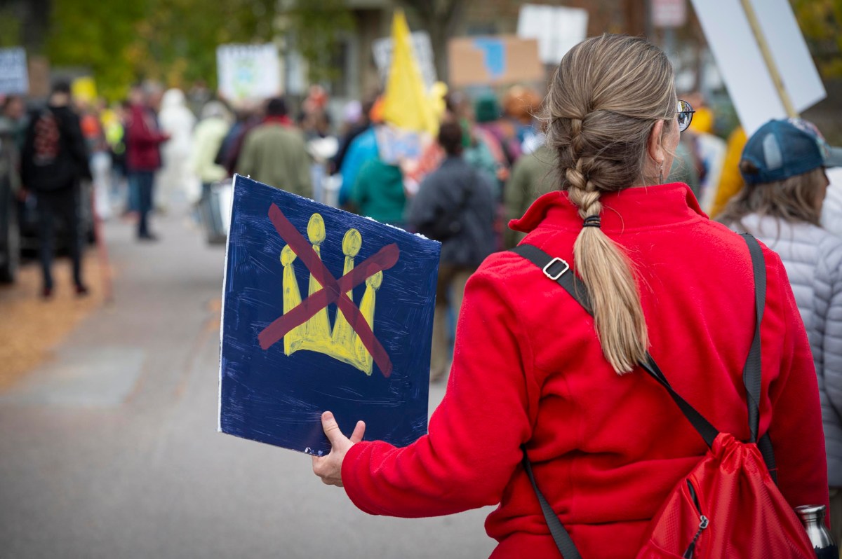 A person in a red coat holds a sign with a yellow crown crossed out at a protest or demonstration, with other participants and signs visible in the background.