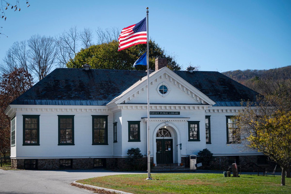 A white, historic public library building with a gabled roof and front entrance, an American flag on a pole in front, and trees in the background on a sunny day.