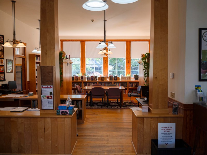 A small library interior with wooden floors, desks, chairs, bookshelves, and large windows letting in natural light.