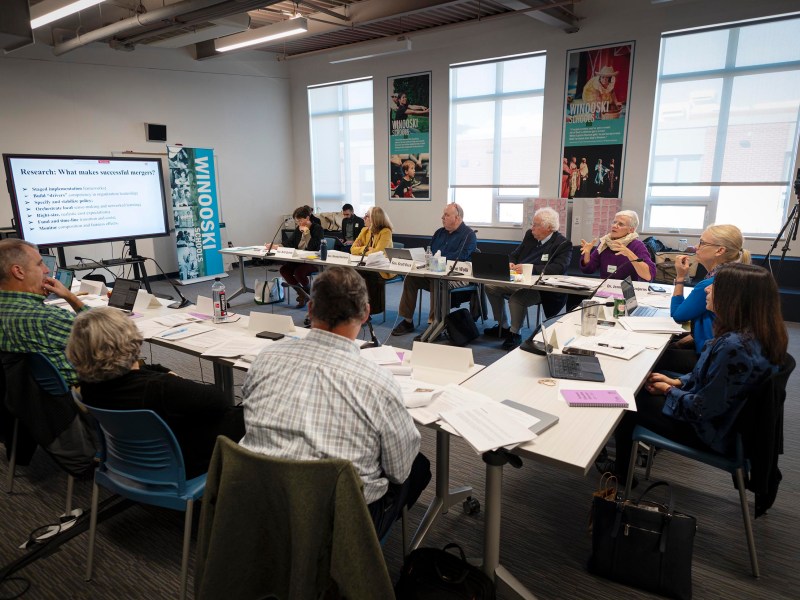 A group of people sits around a conference table in a meeting room, viewing a presentation on a screen titled “Research: What makes successful mergers?”.