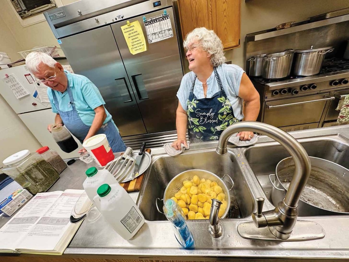 Two people work in a kitchen; one pours liquid while the other smiles at the sink with peeled potatoes soaking in water. Various cooking supplies and a recipe book are visible.