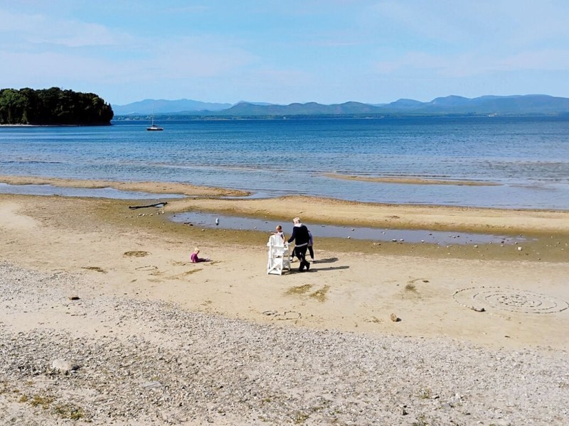 A couple in formal attire walks along a sandy beach near the shoreline, with a child playing nearby and distant mountains in the background.