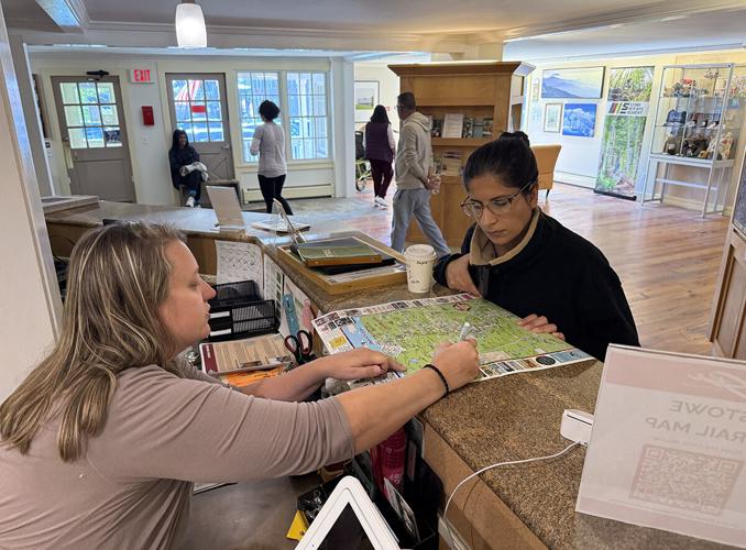 A woman behind a counter points at a map while assisting another woman; several people are in the background of a visitor center.