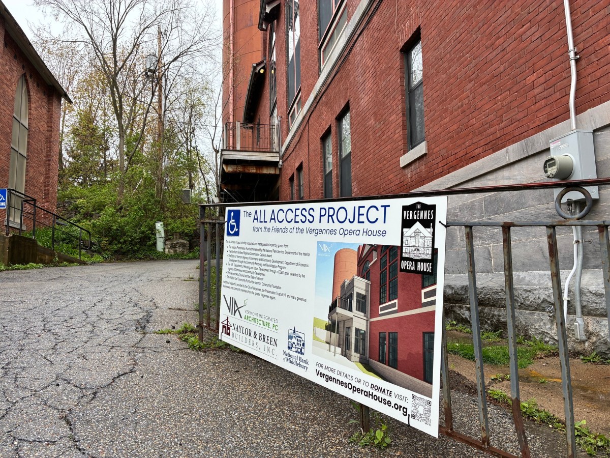 A sign for the All Access Project at Vergennes Opera House is displayed on a railing next to a brick building and paved walkway.