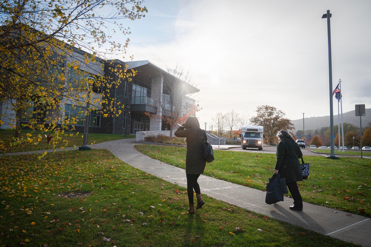 Two people walk on a sidewalk toward a building in the sunlight, carrying bags, with a bus and flagpoles visible in the background.