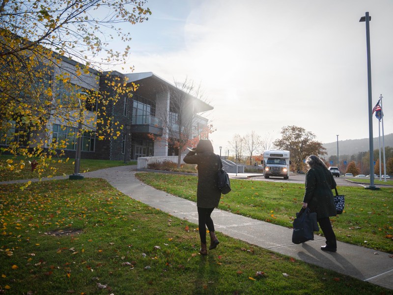 Two people walk on a sidewalk toward a building in the sunlight, carrying bags, with a bus and flagpoles visible in the background.