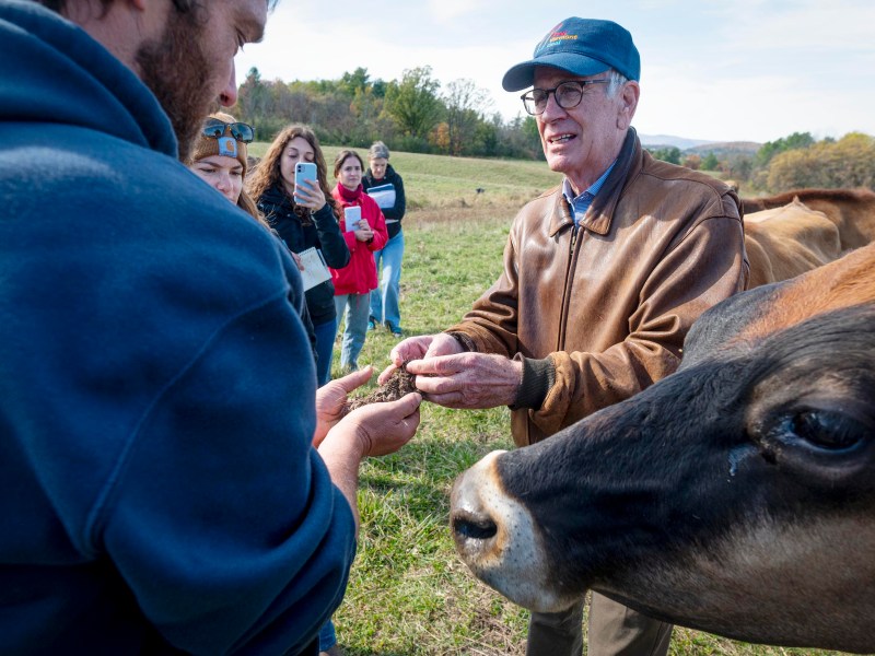 A group of people stand in a field as a man in a leather jacket examines a handful of feed, with a cow in the foreground and others observing and taking photos.