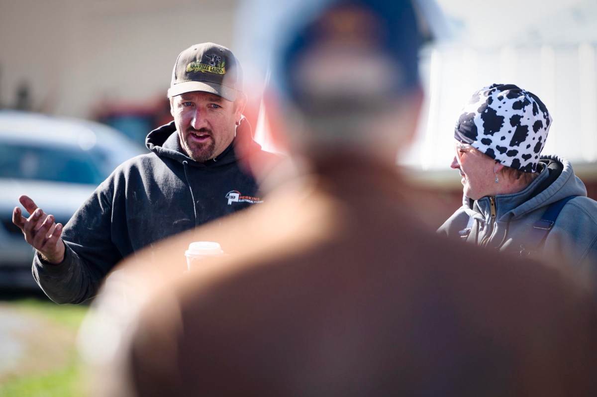 A man wearing a cap gestures while speaking, holding a coffee cup, as another person in a cow-print hat listens; a blurry figure is in the foreground.