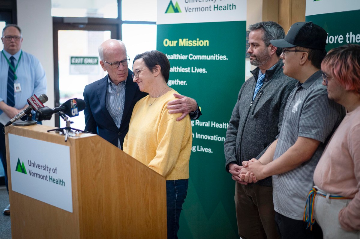 A group of people stands together at a University of Vermont Health press event, with one man comforting a woman at the podium.