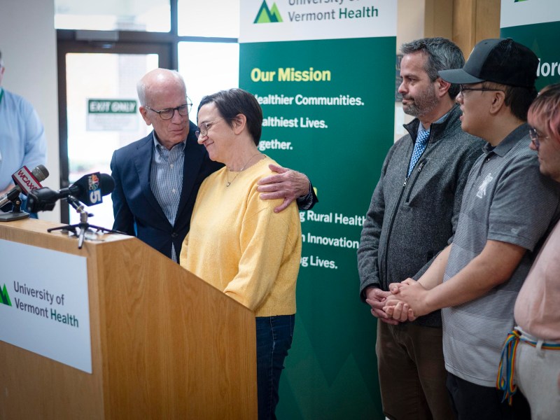 A group of people stands together at a University of Vermont Health press event, with one man comforting a woman at the podium.