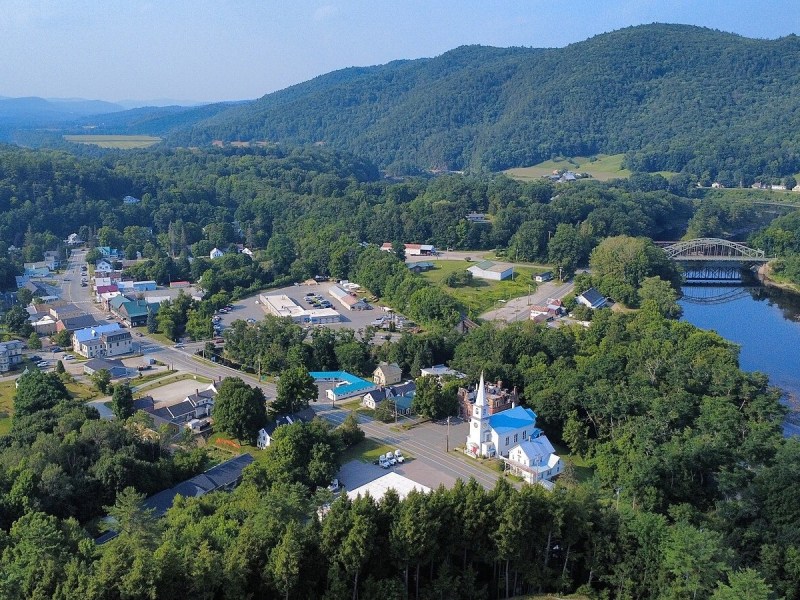 A small town with houses, buildings, a church, and a river sits among green hills and forests under a partly cloudy sky.