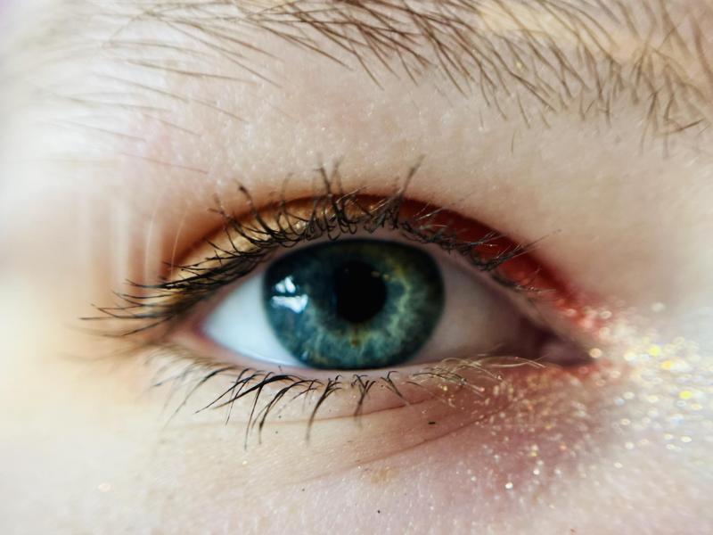 Close-up of a blue eye with long eyelashes, visible eyebrow hairs, and some glitter on the skin below the eye.