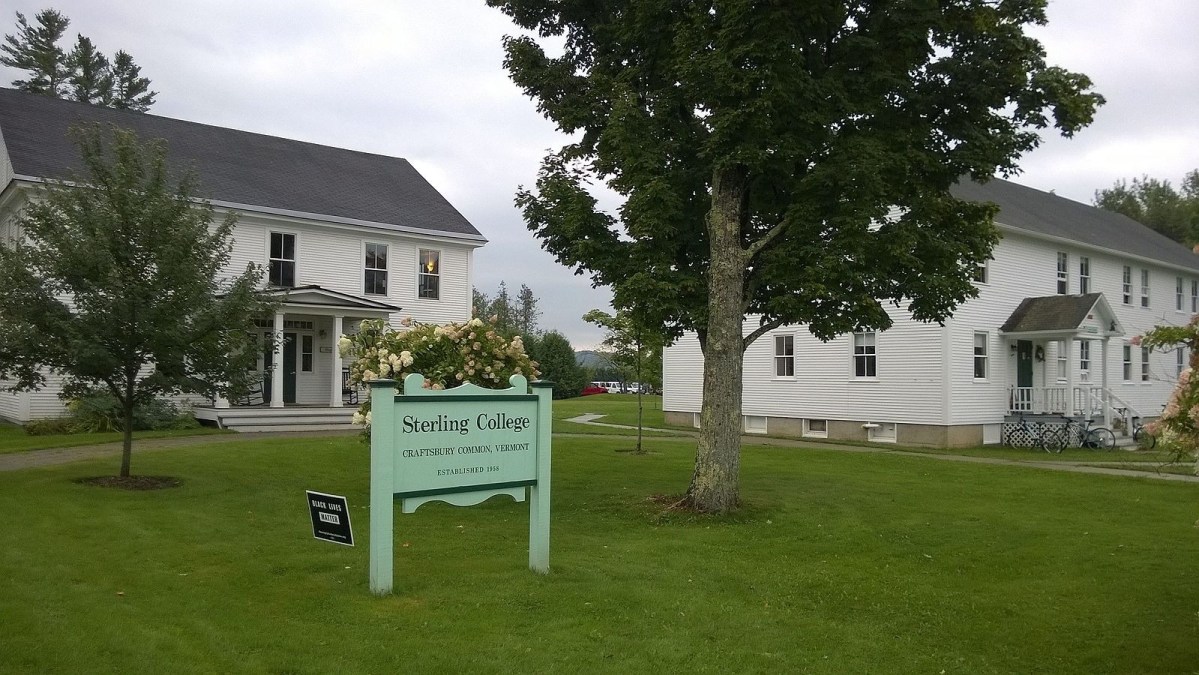 Two white buildings stand on a grassy lawn with trees. A green sign in the foreground reads “Sterling College, Craftsbury Common, Vermont.”.