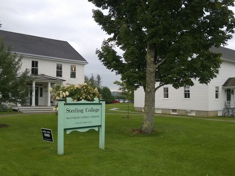 Two white buildings stand on a grassy lawn with trees. A green sign in the foreground reads “Sterling College, Craftsbury Common, Vermont.”.