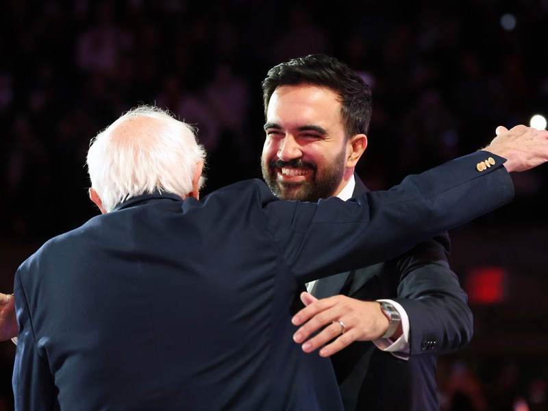 Two men in suits embrace on stage, one facing the camera and smiling, the other with his back to the camera.