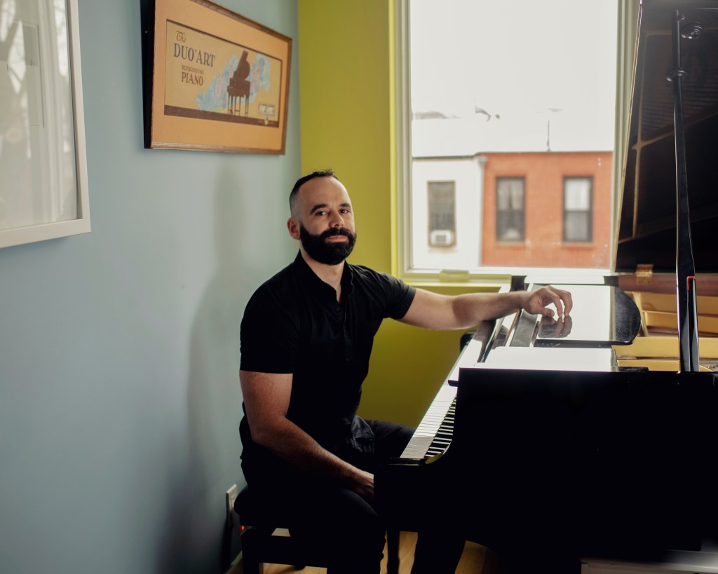 A man with a beard sits at a grand piano in a room with light blue walls and a window showing buildings outside. A framed poster hangs on the wall.