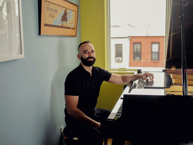 A man with a beard sits at a grand piano in a room with light blue walls and a window showing buildings outside. A framed poster hangs on the wall.