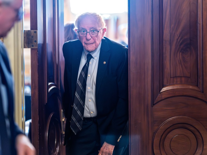 An older man in a suit and tie enters through a large wooden door, with another man partially visible on the left.