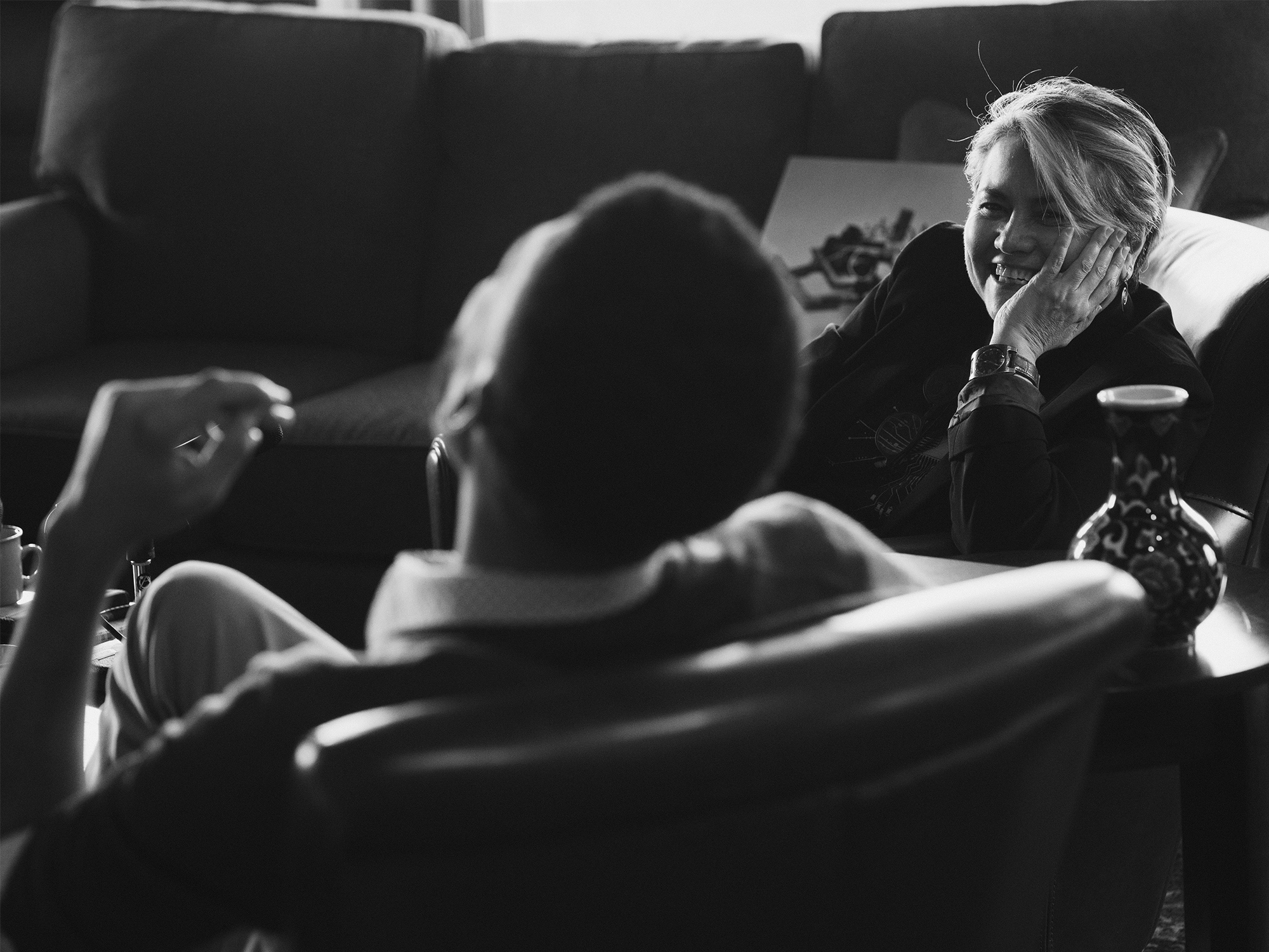 A woman sits on a sofa, smiling with her hand on her face, while another person faces her in conversation in a living room setting.