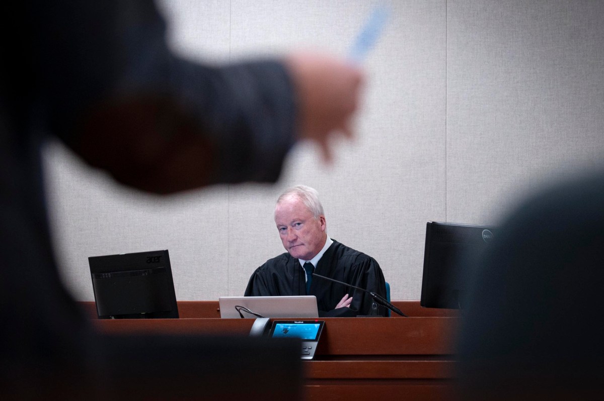 A judge in a black robe sits at a courtroom bench with computers in front of him, looking forward, while a blurred person stands in the foreground.