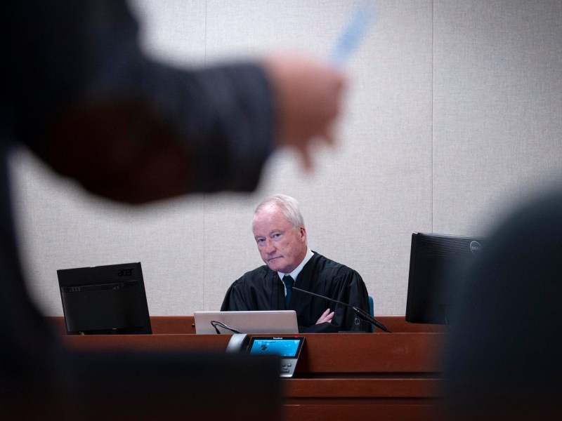 A judge in a black robe sits at a courtroom bench with computers in front of him, looking forward, while a blurred person stands in the foreground.