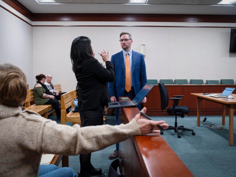 A man in a suit stands facing a woman who is gesturing, while another person in the foreground points toward them in a courtroom setting.