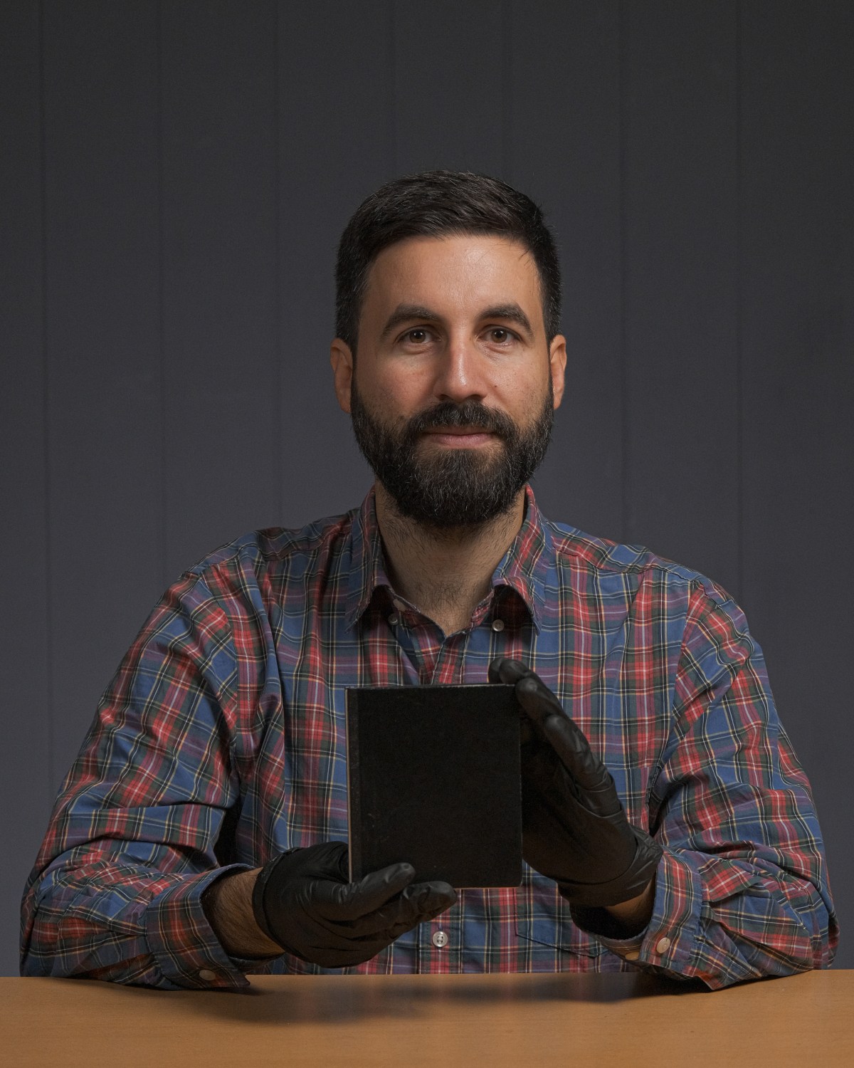 A man with a beard wearing a plaid shirt and black gloves holds a small black rectangular object while sitting at a table against a gray background.
